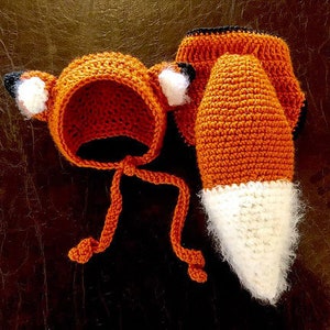 Könnte beinhalten: Ein gehäkeltes Fuchs-Kostüm für ein Baby, bestehend aus einer orangefarbenen Kapuze mit weißen Ohren und einem passenden orange-weißen Schwanz.