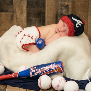 May include: A newborn baby wearing a red, white, and blue baseball cap with the letter "A" on it, and a white and red crocheted diaper with a baseball stitch design. The baby is lying on a white blanket with a blue and red baseball and a blue and red baseball bat with the word "Braves" on it. There are several baseballs scattered around the blanket.