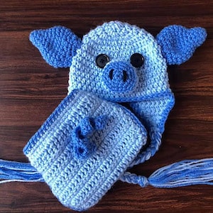 May include: A blue crocheted pig hat with a matching scarf. The hat has two black buttons for eyes and a crocheted snout. The scarf has a long tassel.
