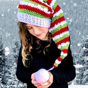 May include: A child wearing a red, white and green striped elf hat with a long, pointed tip. The hat is crocheted and has a pom-pom on the end of the tip. The child is holding a white snowball in their hands.