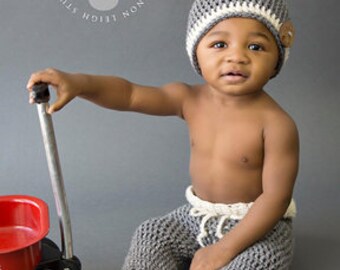 May include: A baby wearing a grey and white crocheted hat and grey crocheted trousers. The baby is holding a red toy wagon.