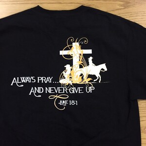 May include: Black t-shirt with a white cross, floral design, and the text "Always Pray... And Never Give Up - Luke 18:1".