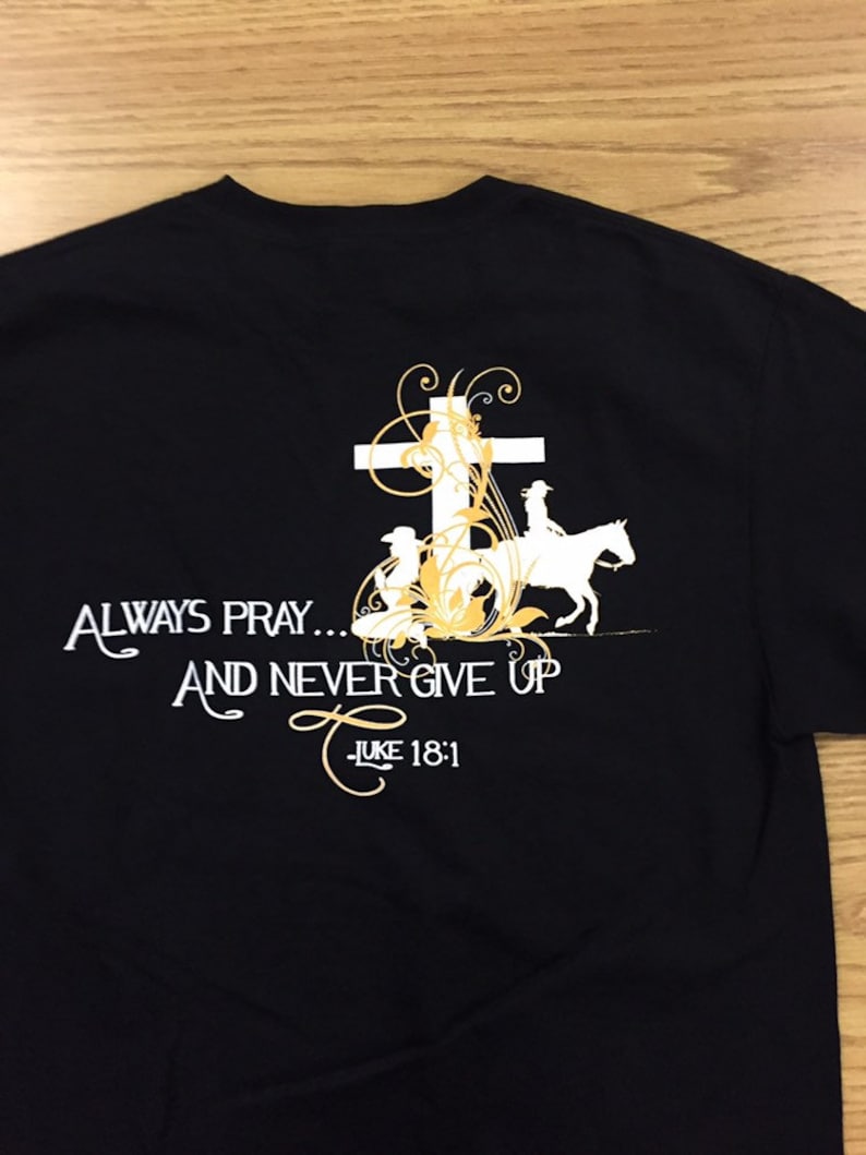 May include: Black t-shirt with a white cross, floral design, and the text "Always Pray... And Never Give Up - Luke 18:1".