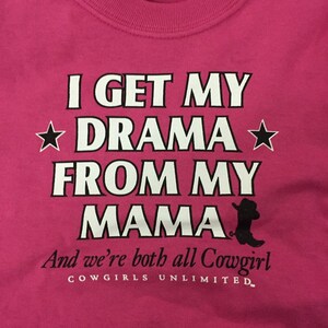 May include: Pink t-shirt with white text that reads "I get my drama from my mama" and a black silhouette of a cowboy boot. The text "And we're both all Cowgirl" is below the boot and the text "COWGIRLS UNLIMITED" is below that.
