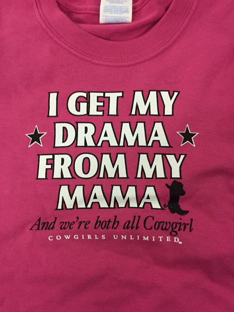 May include: Pink t-shirt with white text that reads "I get my drama from my mama" and a black silhouette of a cowboy boot. The text "And we're both all Cowgirl" is below the boot and the text "COWGIRLS UNLIMITED" is below that.