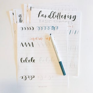 May include: A white sheet of paper with the words "handlettering BASIC STROKES" written in black ink. The paper has several lines of black ink lettering practice, including the words "warm up" and "blele". A gray marker pen is lying on the paper.