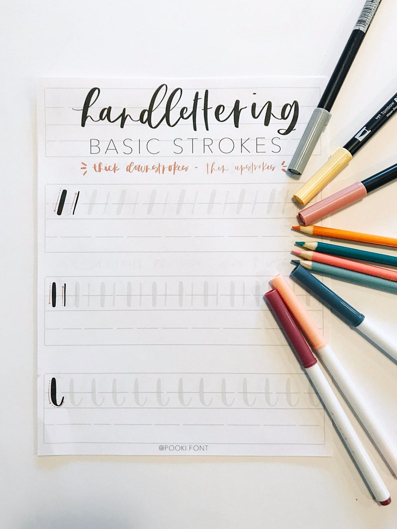 May include: A white sheet of paper with black ink lettering that says "handlettering BASIC STROKES" and shows examples of thick downstrokes and thin upstrokes. The paper is surrounded by various colored markers and pencils.