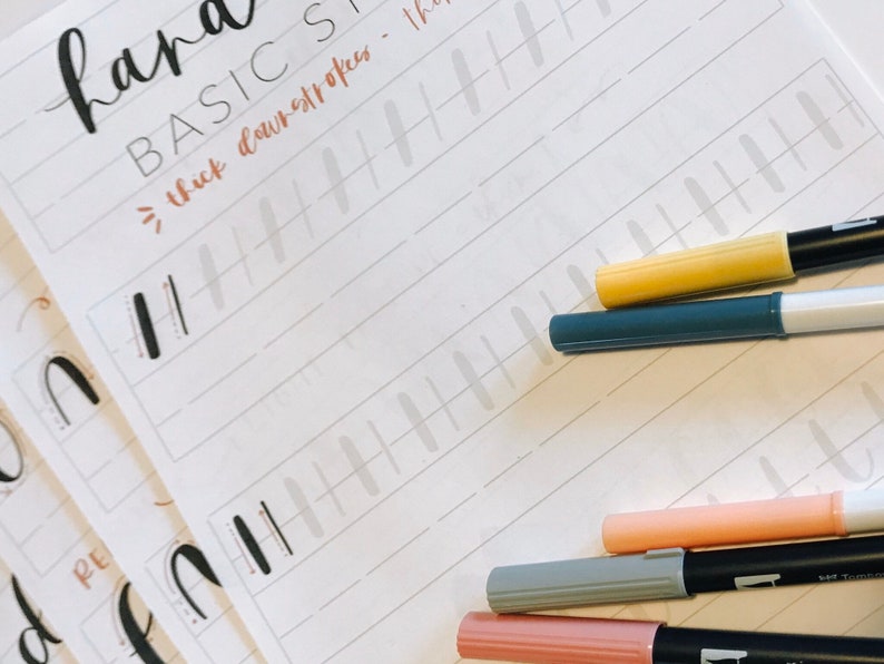 May include: A white sheet of paper with the words "hand lettering basics" and "thick downstrokes" written in black ink. There are several lines of gray lines drawn on the paper. There are five markers on the paper, one yellow, one blue, one pink, one light gray, and one dark gray.