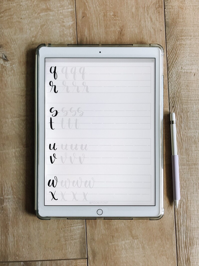 May include: A white iPad with a black screen displaying handwritten letters of the alphabet in cursive. The letters are written in black ink on a white background with gray lines. A purple stylus is lying on a wooden surface next to the iPad.