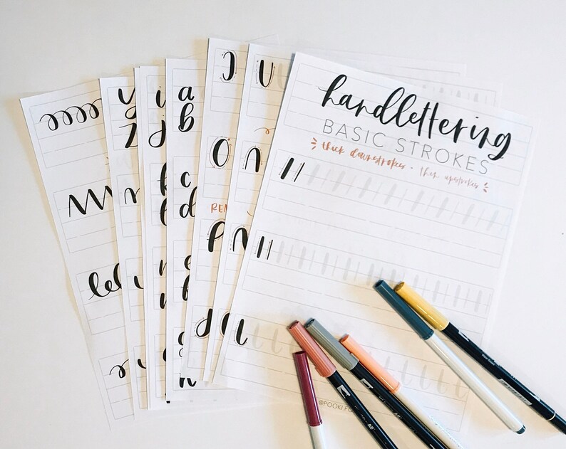 May include: A set of printable hand lettering practice sheets with basic strokes and lowercase letters. The sheets are laid out on a white surface with several colored markers.