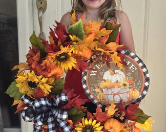 May include: A decorative autumn wreath featuring artificial sunflowers, orange pumpkins, and red and yellow maple leaves. A black and white buffalo check ribbon bow is attached to the side. A round sign in the center reads Welcome with a pumpkin graphic.
