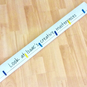 May include: A white wooden sign with the text "Look at Isaac's creative masterpieces" written in black marker. The sign has blue and yellow clothespins attached to it.