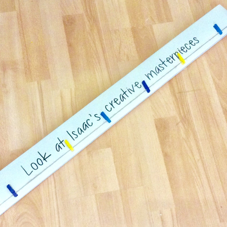 May include: A white wooden sign with the text "Look at Isaac's creative masterpieces" written in black marker. The sign has blue and yellow clothespins attached to it.