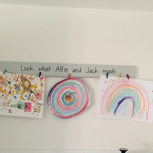 May include: Three colourful drawings by children are displayed on a wooden rack with the text "Look what Alfie and Jack made" written on it.