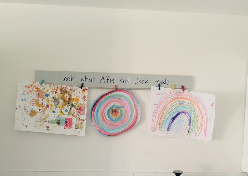 May include: Three colourful drawings by children are displayed on a wooden rack with the text "Look what Alfie and Jack made" written on it.