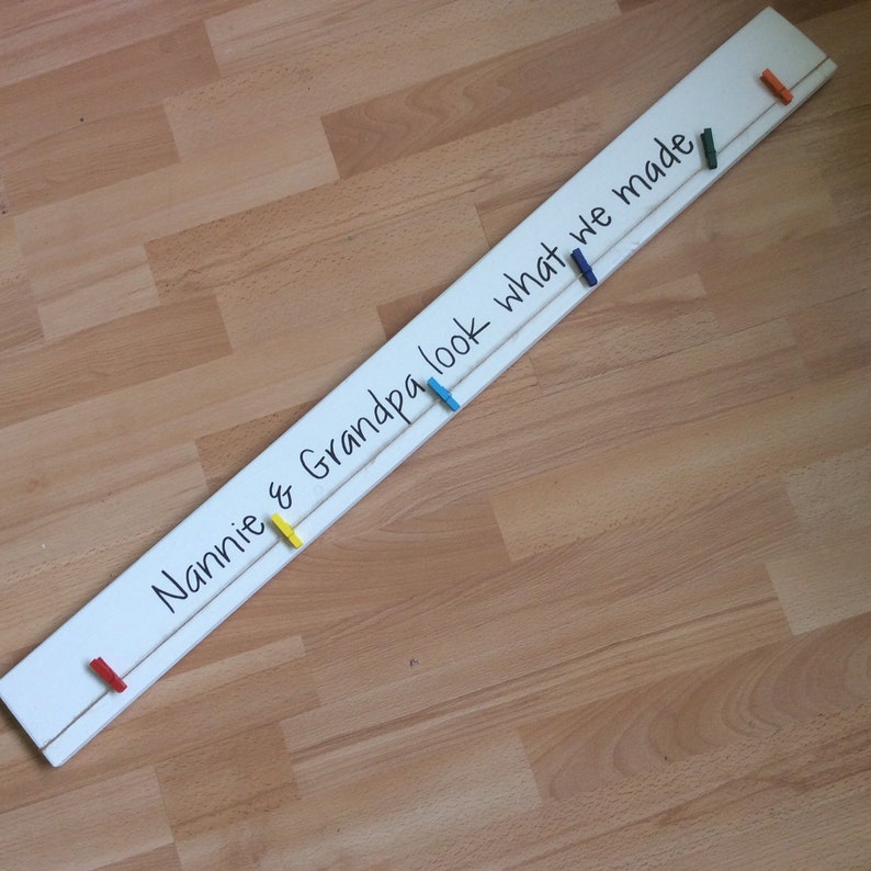 May include: White wooden sign with the text "Nannie & Grandpa look what we made" written in black marker. The sign has a string attached with colourful clothes pegs hanging from it.