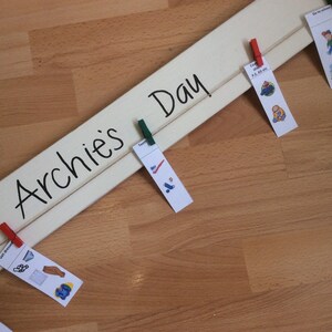 May include: A white wooden board with the text "Archie's Day" written in black.  There are several small cards attached to the board with clothespins. The cards have pictures and text on them, such as "Get Dressed", "Teeth", "Cook", and "Go to School".