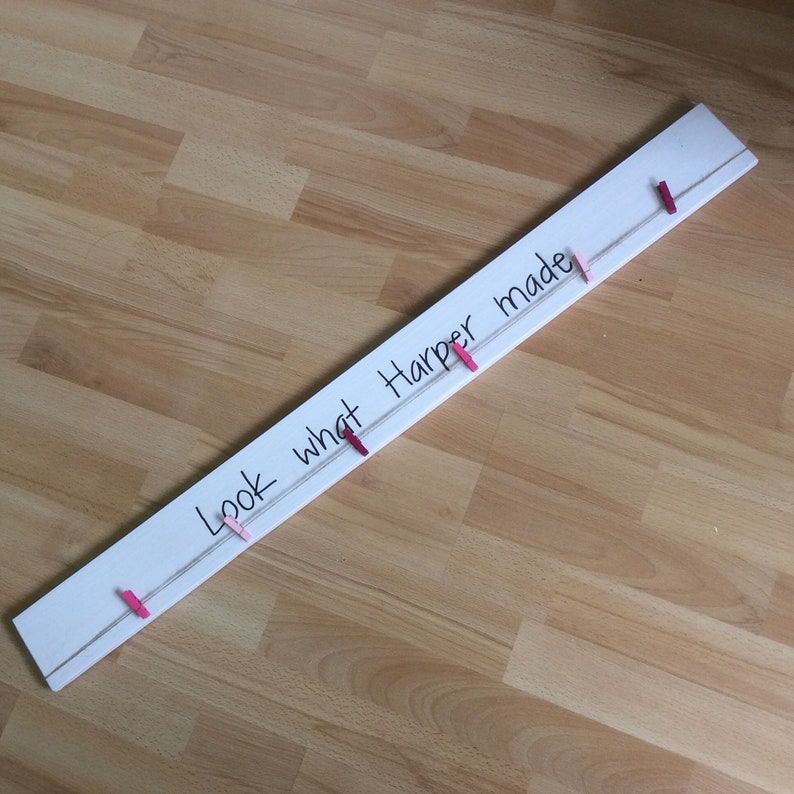 May include: White wooden board with the text "Look what Harper made" written in black marker. A white string is strung across the board with pink and red clothespins attached.
