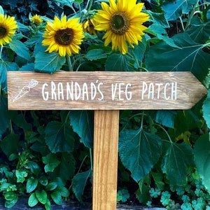 May include: A wooden sign with the text "Grandad's Veg Patch" in white lettering. The sign is propped up on a wooden post and surrounded by green foliage.
