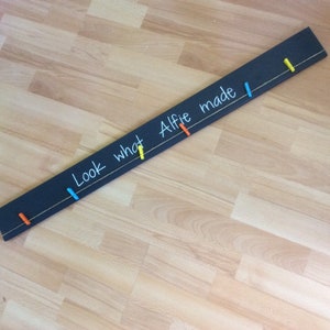 May include: A black chalkboard with the words "Look what Alfie made!" written in white chalk. There are four colourful clothes pegs attached to a string stretched across the board.