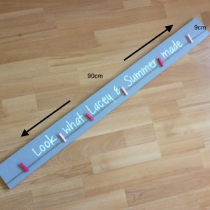 May include: A grey wooden sign with the text "Look what Lacey & Summer made" written in white paint. The sign is 90 cm long and 9 cm wide. It has a string with pink and white clothes pegs attached to it.