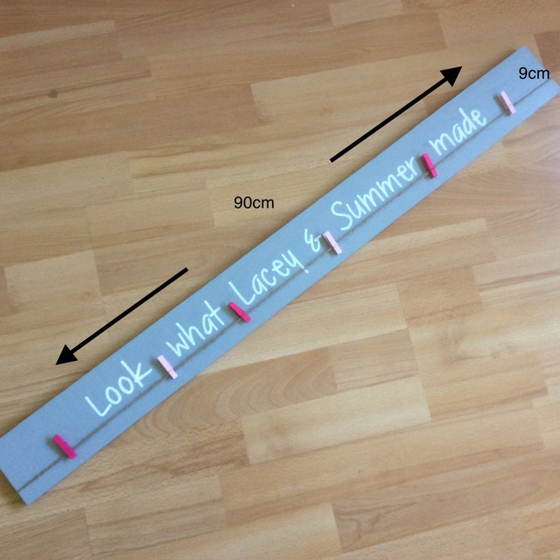 May include: A grey wooden sign with the text "Look what Lacey & Summer made" written in white paint. The sign is 90 cm long and 9 cm wide. It has a string with pink and white clothes pegs attached to it.