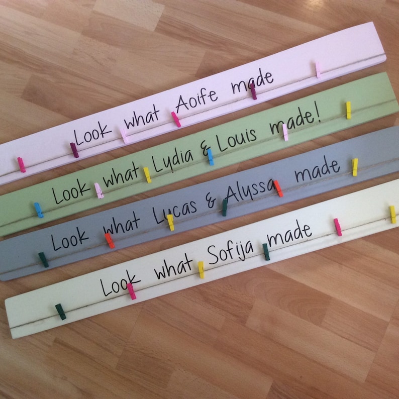May include: Four wooden boards painted in different colours, each with a string and clothes pegs. The boards have handwritten messages that say "Look what Aoife made", "Look what Lydia & Louis made", "Look what Lucas & Alyssa made", and "Look what Sofija made".