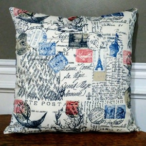 May include: Square decorative pillow with a cream-colored background. The pillow is covered in a collage of vintage postage stamps, handwritten script, and illustrations in shades of blue, red, and black. The Eiffel Tower is visible on the pillow.