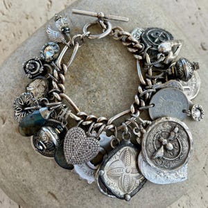 Puede incluir: Una pulsera de plata con cierre de palanca. La pulsera presenta una variedad de dijes, incluyendo monedas, un coraz&oacute;n y otros elementos decorativos. Los dijes est&aacute;n unidos a una pulsera de cadena.