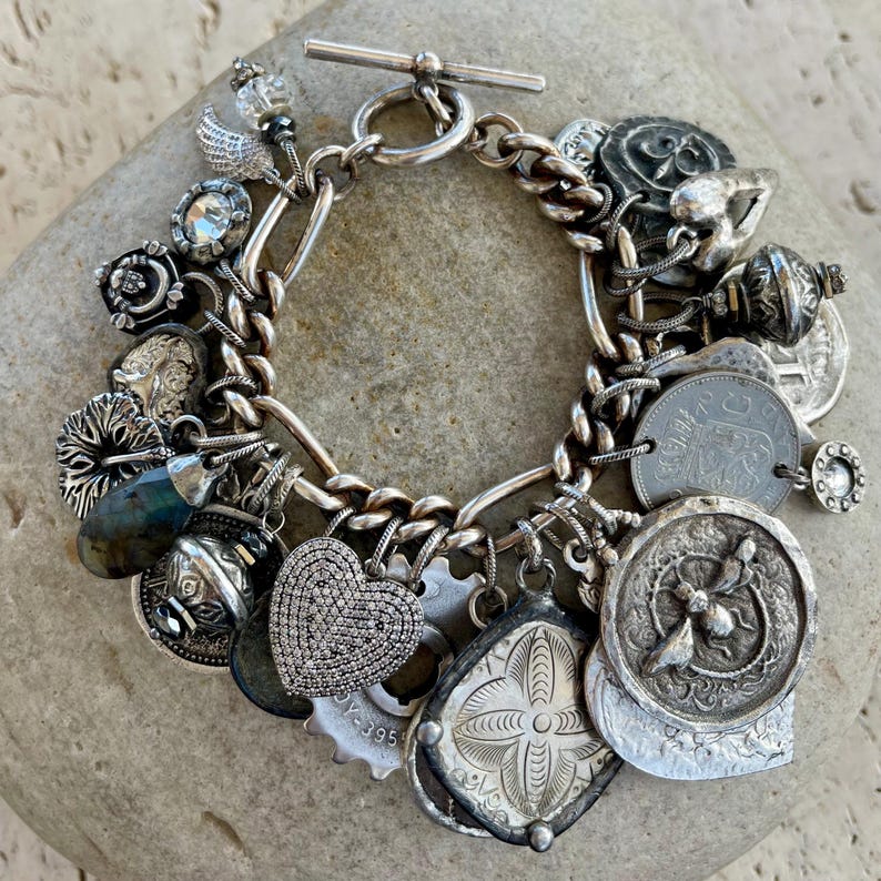 Puede incluir: Una pulsera de plata con cierre de palanca. La pulsera presenta una variedad de dijes, incluyendo monedas, un coraz&oacute;n y otros elementos decorativos. Los dijes est&aacute;n unidos a una pulsera de cadena.