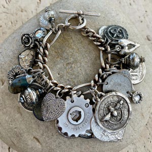 Puede incluir: Una pulsera de plata con cierre de palanca. La pulsera presenta una variedad de dijes, incluyendo monedas, un coraz&oacute;n, un engranaje y otros elementos decorativos. Los dijes est&aacute;n unidos a una pulsera de cadena y cuerda trenzada.