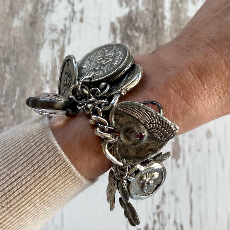 Puede incluir: Pulsera de plata con dijes detallados, que incluyen colgantes en forma de moneda, un dije en forma de coraz&oacute;n con una piedra roja y dijes en forma de pluma. La pulsera est&aacute; hecha de cadenas de plata y se usa en la mu&ntilde;eca.