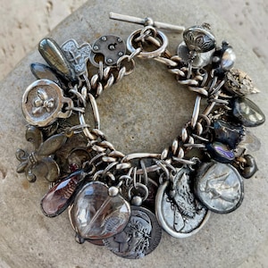 Puede incluir: Una pulsera de plata con dijes que presenta una variedad de abalorios, incluyendo monedas, medallones y colgantes en forma de coraz&oacute;n. La pulsera est&aacute; hecha de una cadena gruesa y tiene un cierre de palanca. Los dijes son detallados y tienen un aspecto antiguo.