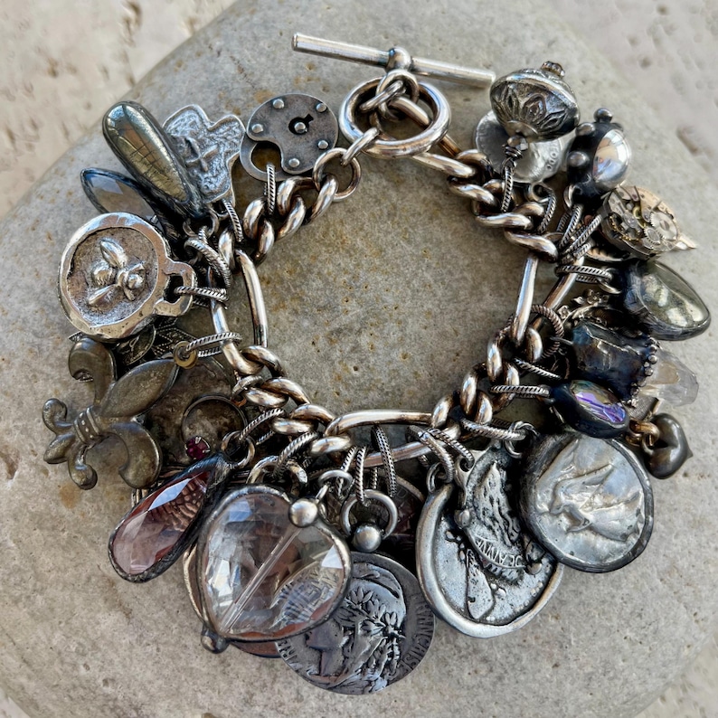 Puede incluir: Una pulsera de plata con dijes que presenta una variedad de abalorios, incluyendo monedas, medallones y colgantes en forma de coraz&oacute;n. La pulsera est&aacute; hecha de una cadena gruesa y tiene un cierre de palanca. Los dijes son detallados y tienen un aspecto antiguo.