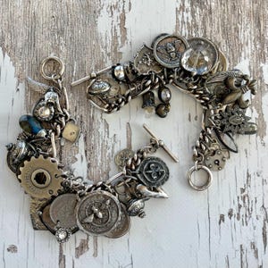 Puede incluir: Dos pulseras de plata con dijes de estilo vintage, que incluyen monedas, engranajes y medallones. Las pulseras est&aacute;n conectadas por una cadena y un cierre de palanca, sobre una superficie de madera blanca envejecida.