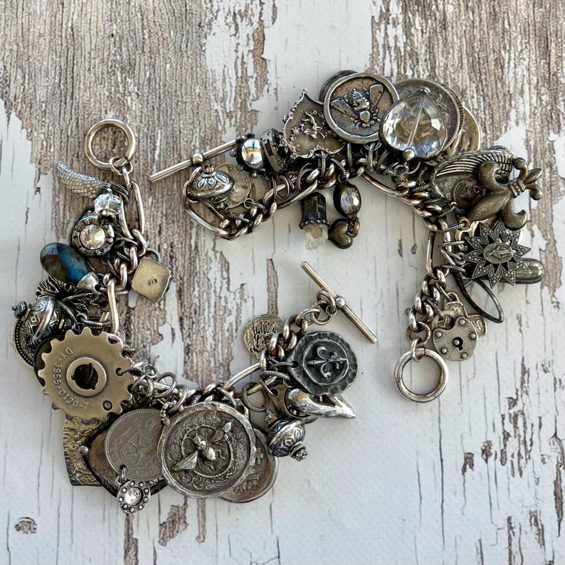 Puede incluir: Dos pulseras de plata con dijes de estilo vintage, que incluyen monedas, engranajes y medallones. Las pulseras est&aacute;n conectadas por una cadena y un cierre de palanca, sobre una superficie de madera blanca envejecida.