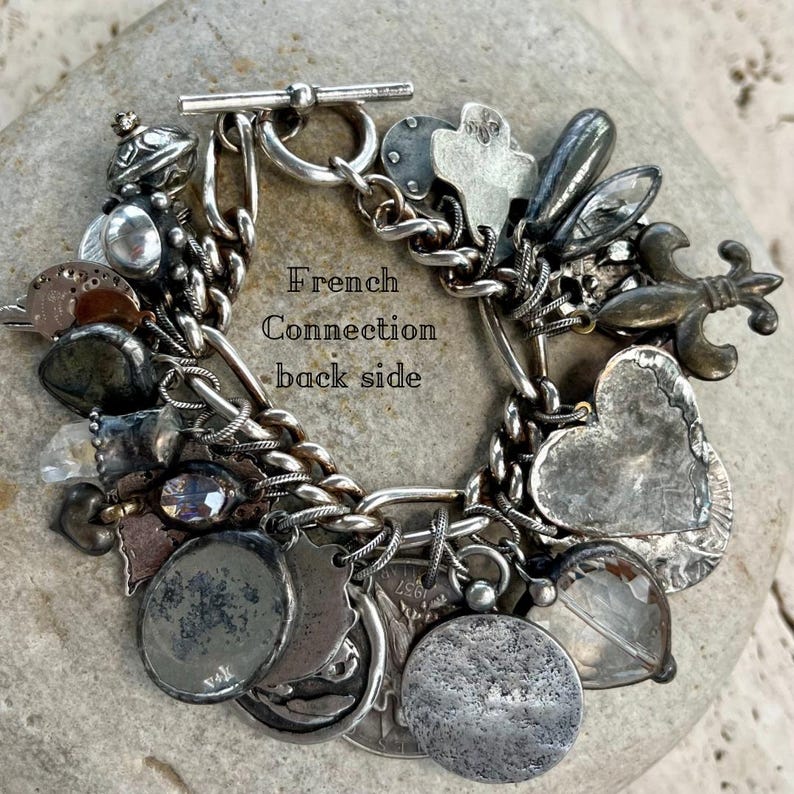 Puede incluir: Pulsera de abalorios de plata con cierre de palanca, con varios abalorios que incluyen corazones, monedas y una flor de lis. La pulsera se muestra sobre una superficie de piedra. El texto de la pulsera dice "French Connection back side."