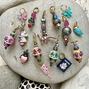 May include: A collection of colorful charms, including a unicorn, dinosaur, skull, lightning bolt, butterfly, and a passport. The charms are made of various materials, including ceramic, metal, and glass. They are strung on gold chains and are perfect for adding a touch of whimsy to any jewelry project.