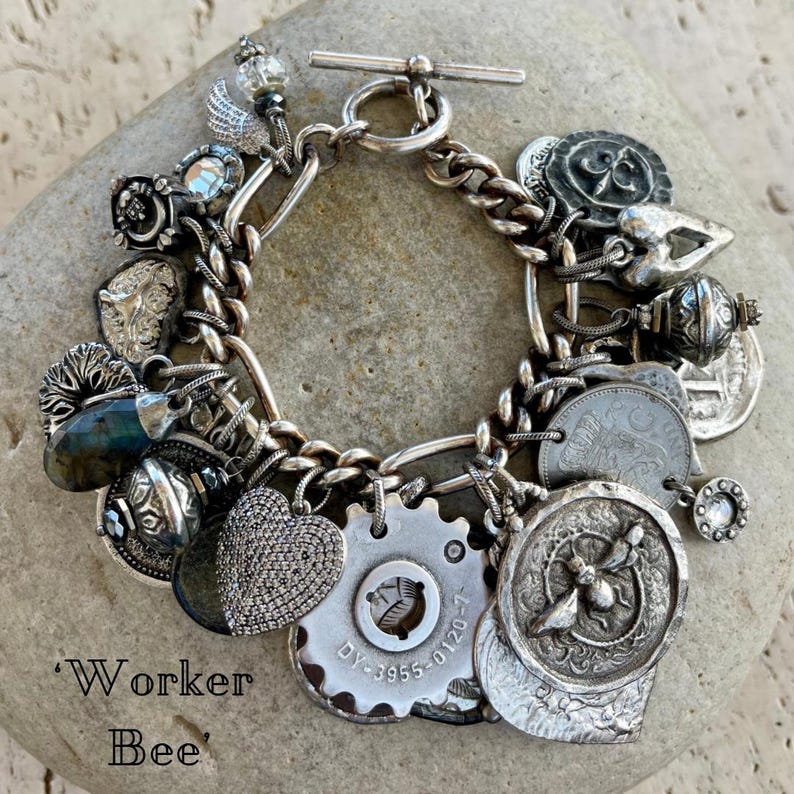 Puede incluir: Pulsera de plata con dijes variados, incluyendo un coraz&oacute;n, monedas, un engranaje y una abeja. La pulsera tiene un cierre de palanca y se muestra sobre una piedra. El texto "Worker Bee" es visible.