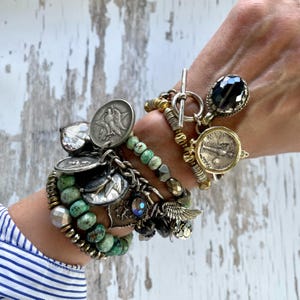 Puede incluir: Una colecci&oacute;n de pulseras superpuestas con varios dijes y cuentas. Las pulseras presentan monedas de plata, cristales facetados, cuentas turquesas y detalles met&aacute;licos. Los estilos incluyen cierres de palanca y una mezcla de texturas y colores.