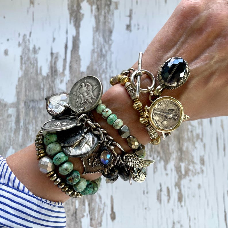 Puede incluir: Una colecci&oacute;n de pulseras superpuestas con varios dijes y cuentas. Las pulseras presentan monedas de plata, cristales facetados, cuentas turquesas y detalles met&aacute;licos. Los estilos incluyen cierres de palanca y una mezcla de texturas y colores.