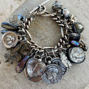 Puede incluir: Pulsera de plata con dijes y cierre de palanca. La pulsera presenta varios dijes, incluyendo monedas, una flor de lis y otros elementos decorativos. Los dijes est&aacute;n unidos a una pulsera de cadena.