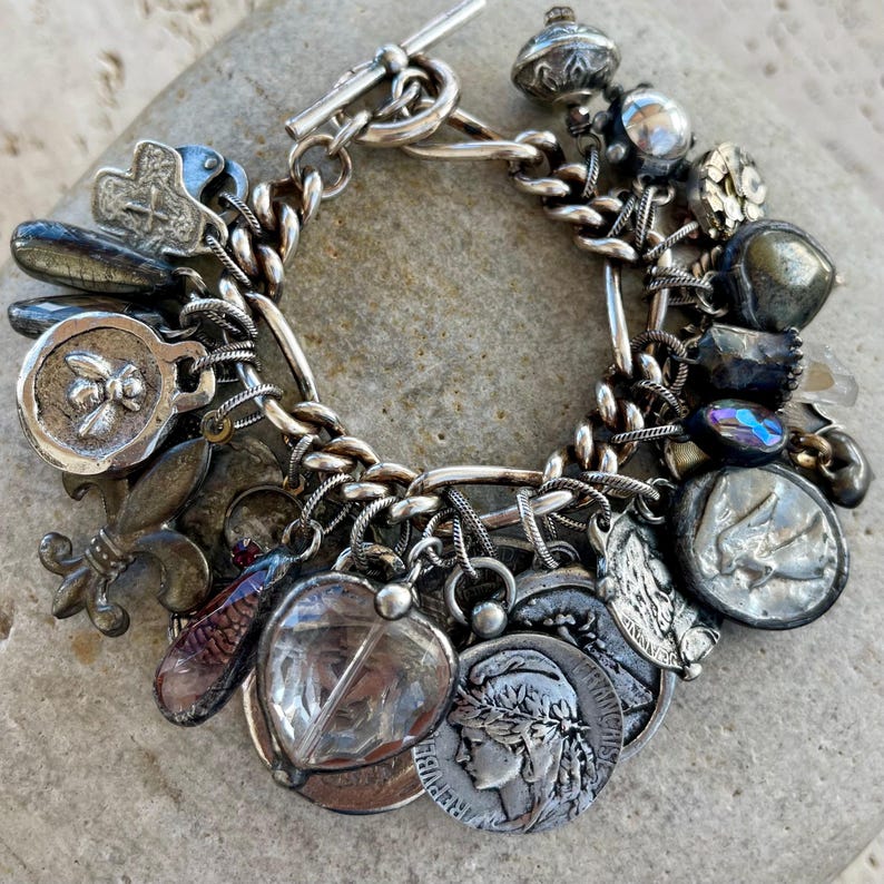 Puede incluir: Pulsera de plata con dijes y cierre de palanca. La pulsera presenta varios dijes, incluyendo monedas, una flor de lis y otros elementos decorativos. Los dijes est&aacute;n unidos a una pulsera de cadena.