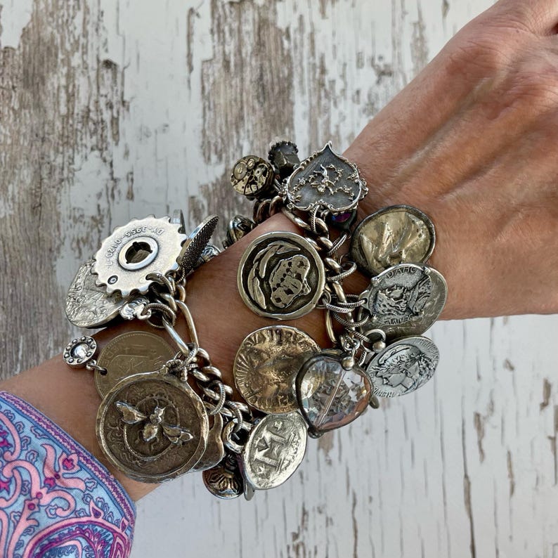 Puede incluir: Una pulsera de plata con una variedad de dijes vintage, incluyendo monedas, engranajes y colgantes en forma de coraz&oacute;n. La pulsera est&aacute; hecha de cadenas enlazadas y tiene una est&eacute;tica r&uacute;stica y antigua.