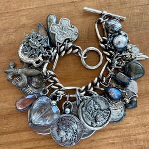 Puede incluir: Pulsera de plata con dijes variados, incluyendo monedas, una cruz, una estrella y un dije en forma de coraz&oacute;n. La pulsera tiene un cierre de palanca y una cadena gruesa. Los dijes son detallados y tienen un aspecto antiguo.