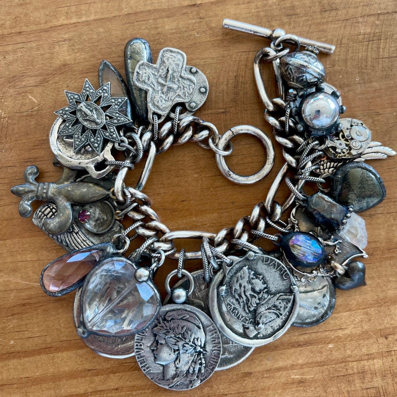Puede incluir: Pulsera de plata con dijes variados, incluyendo monedas, una cruz, una estrella y un dije en forma de coraz&oacute;n. La pulsera tiene un cierre de palanca y una cadena gruesa. Los dijes son detallados y tienen un aspecto antiguo.