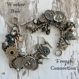 Puede incluir: Dos pulseras de plata con dijes de estilo vintage, incluyendo monedas, engranajes y medallones. Una pulsera est&aacute; etiquetada como 'Worker Bee', y la otra como 'French Connection'. Se exhiben sobre una superficie de madera blanca desgastada.