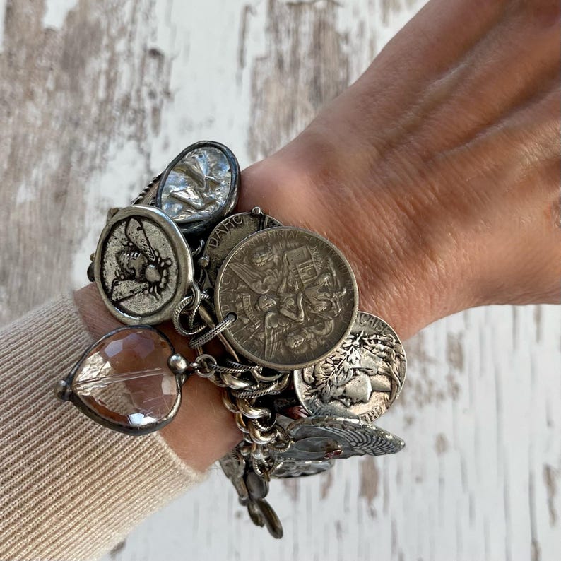 Puede incluir: Una pulsera de plata con dijes variados, incluyendo monedas, una abeja y un cristal en forma de coraz&oacute;n. La pulsera se lleva en la mu&ntilde;eca, mostrando los dise&ntilde;os y texturas detalladas de los dijes. La pulsera es de metal plateado.
