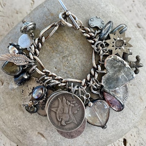 Puede incluir: Pulsera de plata con dijes y cierre de palanca, adornada con diversos dijes como monedas, corazones y estrellas. La pulsera se exhibe sobre una piedra gris. Los dijes muestran dise&ntilde;os detallados y texturas variadas.