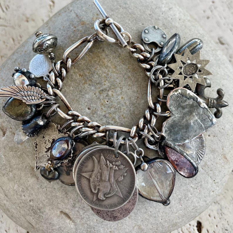 Puede incluir: Pulsera de plata con dijes y cierre de palanca, adornada con diversos dijes como monedas, corazones y estrellas. La pulsera se exhibe sobre una piedra gris. Los dijes muestran dise&ntilde;os detallados y texturas variadas.